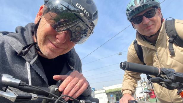 Zwei Männer mit Fahrradhelmen und Sonnenbrillen stehen mit ihren Fahrrädern an einer Straßenkreuzung bei sonnigem Wetter.