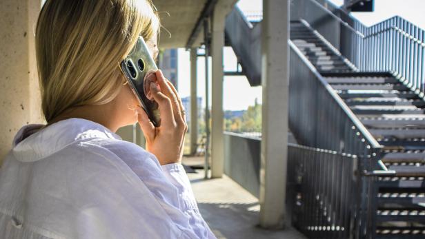 Eine Frau mit blondem Haar telefoniert draußen neben einer Treppe und trägt ein weißes Hemd.