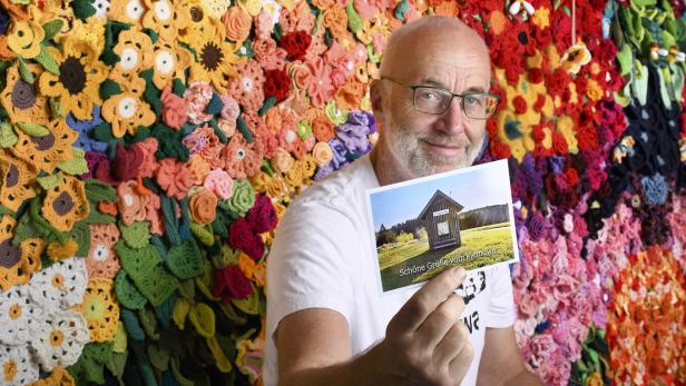 Ein älterer Mann mit Brille hält eine Postkarte vor einer bunten Wand aus gehäkelten Blumenmustern.