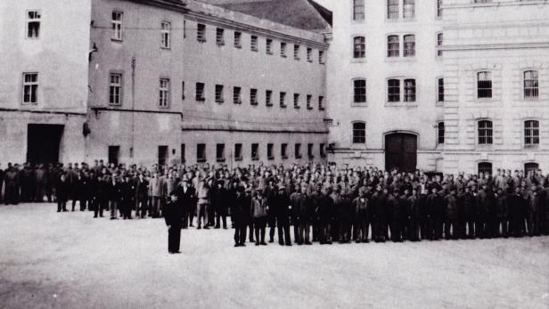 Viele Menschen stehen in mehreren Reihen auf einem Hof vor großen, mehrstöckigen Gebäuden mit vielen Fenstern.