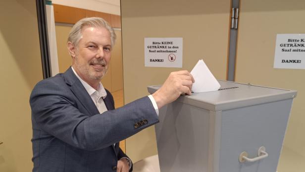 Ein Mann im Anzug wirft einen Wahlzettel in eine graue Wahlurne und lächelt dabei.