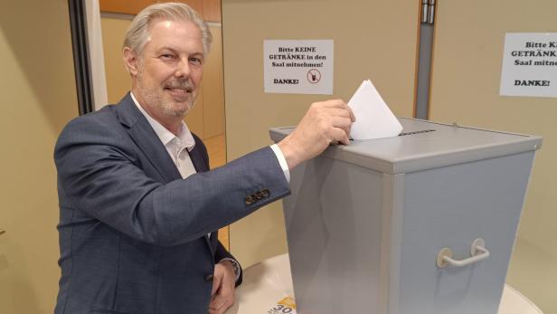 Ein Mann im Anzug wirft einen Wahlzettel in eine graue Wahlurne und lächelt dabei.