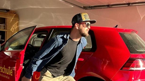 Ein Mann mit Sonnenbrille und Kappe steigt eilig aus einem roten Auto in einer Garage.