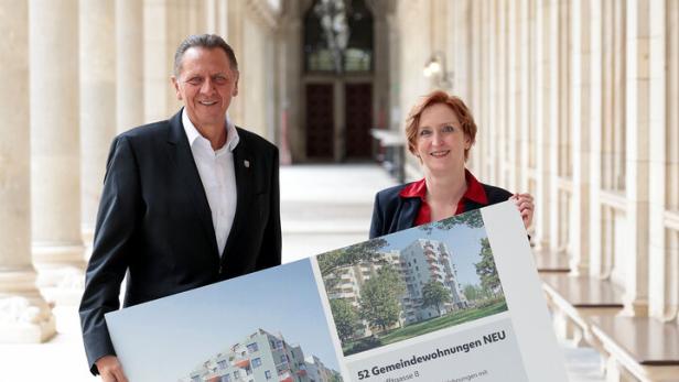 Zwei Personen in Businesskleidung halten ein großes Plakat mit Informationen zu neuen Gemeindewohnungen in einem Arkadengang.