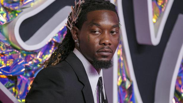 Offset, mit Zöpfen und Anzug, trägt ein silbernes Haaraccessoire vor einem schillernden Hintergrund.