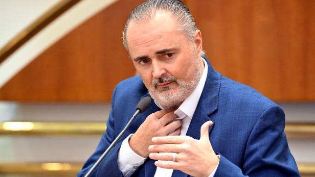 Ein Mann im blauen Anzug spricht gestikulierend an einem Mikrofon in einem Innenraum mit Holzverkleidung.