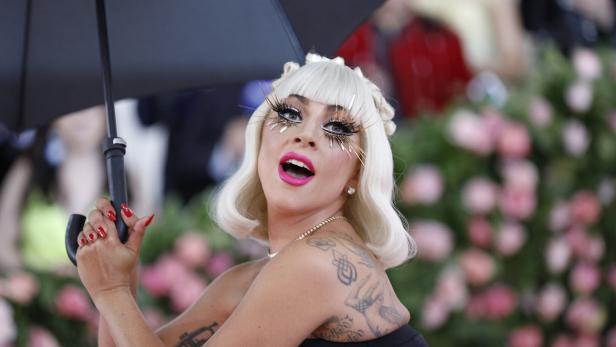 Lady Gaga mit weißblonder Perücke, auffälligem Make-up und Tattoos hält einen Regenschirm.