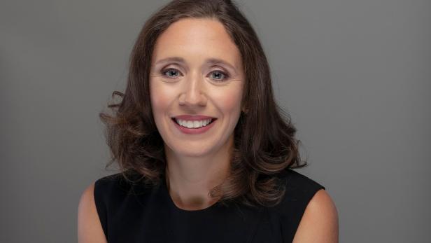 Frau mit schulterlangem braunem Haar und schwarzem ärmellosem Oberteil lächelt vor grauem Hintergrund.