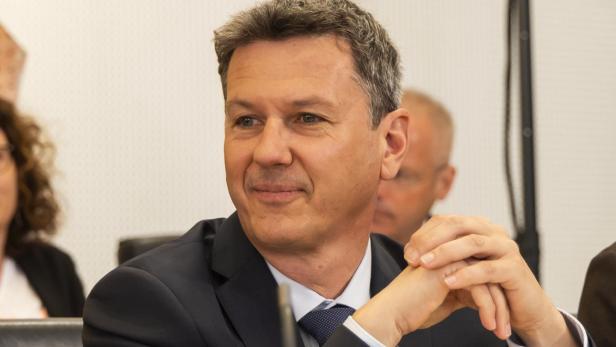 Ein Mann im Anzug sitzt an einem Konferenztisch mit Mikrofon und Wasserglas und lächelt leicht.