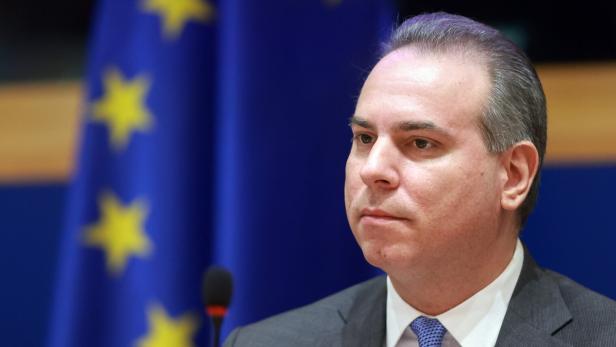 Ein Mann im Anzug sitzt vor einer EU-Flagge und einem Mikrofon.