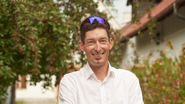 Ein Mann mit weißem Hemd und Sonnenbrille auf dem Kopf steht lächelnd mit verschränkten Armen auf einem gepflasterten Weg im Grünen.