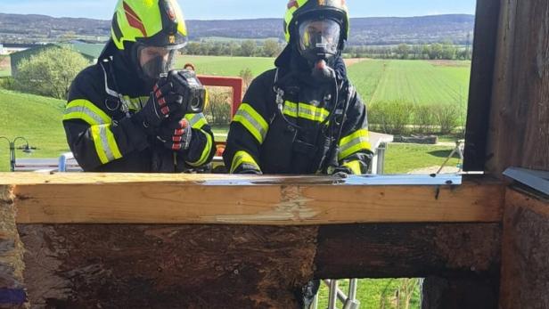Zwei Feuerwehrleute in Schutzkleidung und Atemschutzmasken stehen an einer verbrannten Holzwand und benutzen eine Wärmebildkamera.