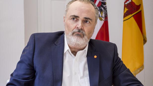 Ein Mann im Anzug sitzt an einem Tisch mit einem Glas Wasser und einem Smartphone, im Hintergrund zwei Fahnen.