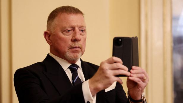 Boris Becker im Anzug hält ein Smartphone hoch.