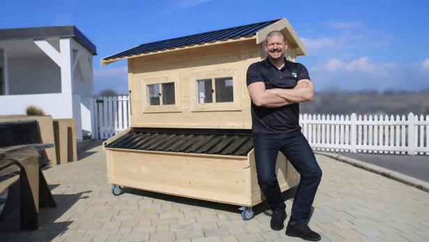 Mann steht lächelnd vor einem großen, mobilen Hühnerstall aus Holz im Freien.