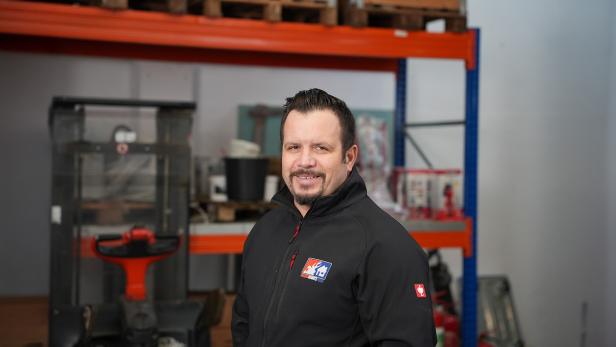 Mann mit schwarzer Jacke steht in einem Lager mit Regalen und Arbeitsmaterialien im Hintergrund.