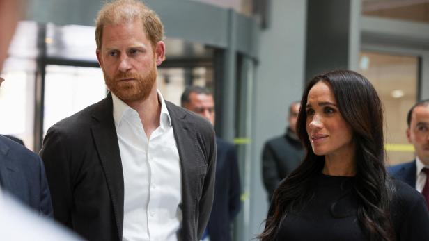 Prinz Harry und Herzogin Meghan, in formeller Kleidung, stehen in einem hellen Innenraum.