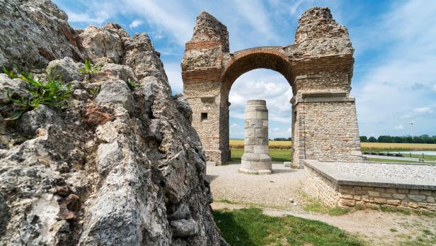Eine alte Steinruine mit einem Bogen steht vor einem Feld unter blauem Himmel.
