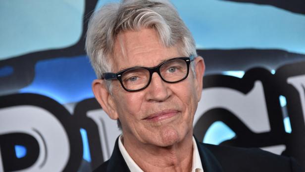 Eric Roberts mit grauen Haaren und Brille lächelt vor einem blauen Hintergrund.