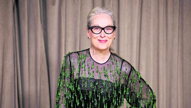 Meryl Streep lächelt mit Brille und grün verziertem, schwarzem Oberteil vor beigem Hintergrund.