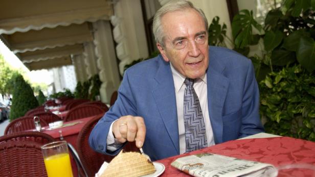 Ein älterer Mann im blauen Anzug sitzt an einem Tisch im Freien, isst eine Waffel und hat ein Glas Orangensaft vor sich.