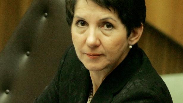 Eine Frau mit kurzen dunklen Haaren und schwarzem Blazer sitzt an einem Tisch und blickt ernst in die Kamera.