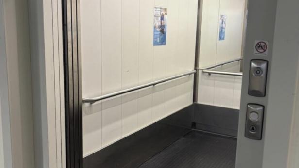 Ein offener Aufzug mit Spiegelwand, Haltegriff und schwarzem Boden ist zu sehen.