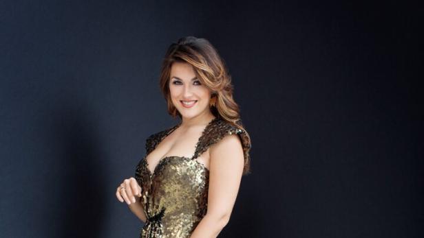 Frau mit langen Haaren trägt ein funkelndes, bodenlanges, goldenes Abendkleid mit Beinschlitz und lächelt.