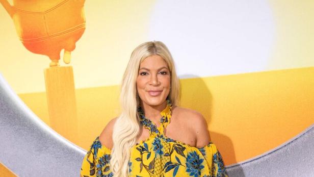 Tori Spelling mit blonden Haaren trägt ein gelbes, geblümtes Kleid vor einem gelben Hintergrund.