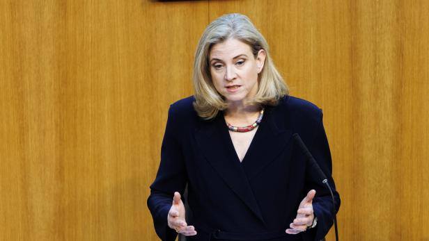 Eine Frau mit blondem Haar spricht gestikulierend an einem Rednerpult vor einer holzvertäfelten Wand.