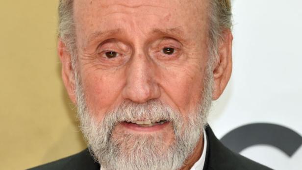 Ray Stevens mit grauem Bart und Anzug, dessen Einstecktuch orange ist, blickt in die Kamera.