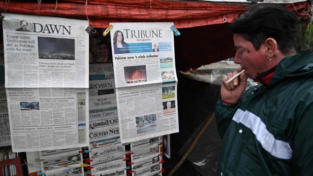 Eine Person mit Zigarette betrachtet eine Auswahl an Zeitungen, die an einem Straßenstand aufgehängt sind.