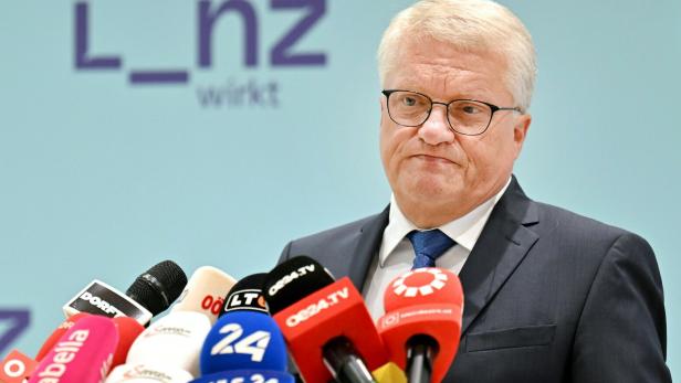 Ein Mann im Anzug steht vor mehreren Mikrofonen und spricht mit ernstem Gesichtsausdruck.