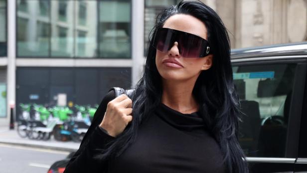 Katie Price mit langen schwarzen Haaren und großer Sonnenbrille trägt schwarzes Oberteil und steht auf einer Straße vor einem Auto.