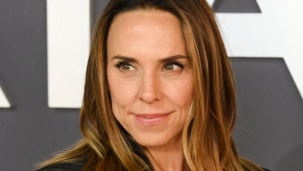 Melanie Chisholm lächelt mit offenem Haar und schwarzem Blazer vor einem grauen Hintergrund.