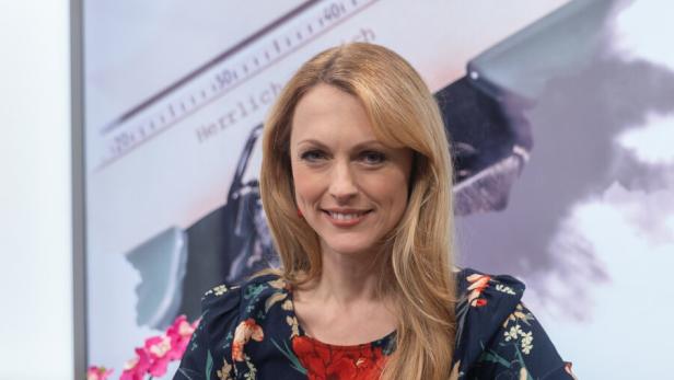 Eine Frau mit langen blonden Haaren sitzt lächelnd auf einem roten Sessel, im Hintergrund sind Blumen und ein Buch zu sehen.