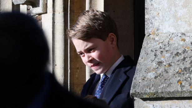 Prinz George mit Anzug und Krawatte steht vor einer steinernen Wand.