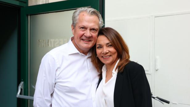 Ein lächelndes Paar, er im weißen Hemd, sie im Blazer, posiert vor einer Tür.