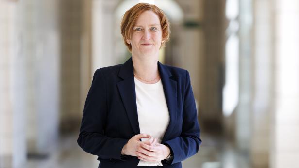 Elke Habel-Torsch mit kurzem, rötlichem Haar trägt einen dunkelblauen Anzug und steht in einem hellen Flur mit Säulen im Wiener Rathaus.