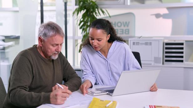 Eine Frau berät einen Mann bei Dokumenten am Schreibtisch mit Laptop und Taschenrechner.