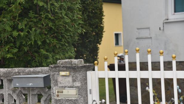 Weiße Gartentür mit goldenen Kugeln, grauer Briefkasten und Klingel an einer Steinmauer, dahinter grüne Hecke und gelbes Haus.
