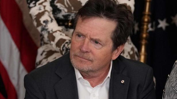 Michael J. Fox sitzt im Anzug, die Hände gefaltet, im Hintergrund Flaggen und Personen.