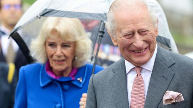 König Charles III. lächelt unter einem Regenschirm, neben ihm Königin Camilla in einem blauen Mantel.