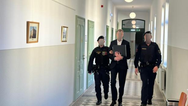 Ein Mann in Anzug wird von zwei Polizisten durch einen hellen Flur begleitet.