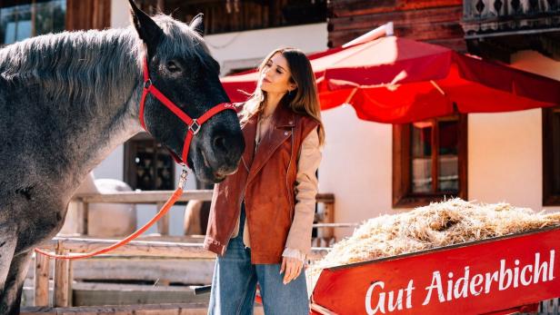 Eine Frau steht neben einem grauen Pferd mit rotem Halfter und einer roten Schubkarre voller Stroh mit der Aufschrift „Gut Aiderbichl“.