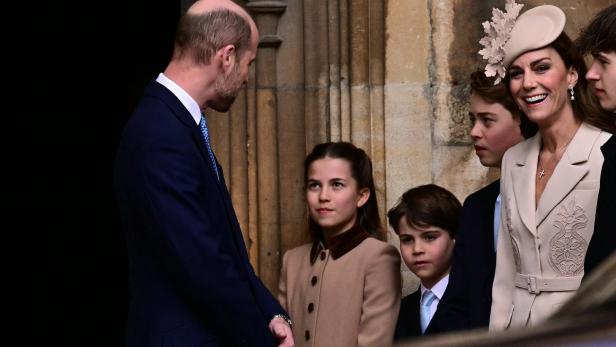 Mehrere elegant gekleidete Personen, darunter Prinz William, Prinzessin Kate und ihre Kinder George, Charlotte und Louis, stehen vor einem steinernen Gebäude und unterhalten sich, eine Frau trägt einen auffälligen Hut.
