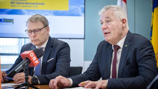 Zwei Männer in Anzügen sitzen an einem Tisch mit Mikrofonen und Flaggen im Hintergrund.