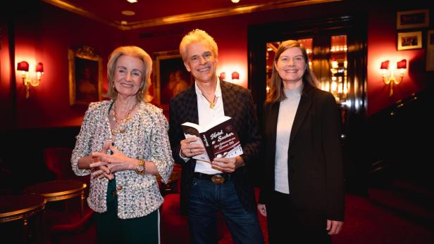 Drei Personen stehen nebeineinader vor dunklem Hintergrund, Thomas Brezina hält ein Buch in Händen