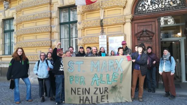 Eine Gruppe von Menschen steht vor einem historischen Gebäude und hält ein Transparent mit der Aufschrift „St. Marx für alle! Nein zur Halle!“.