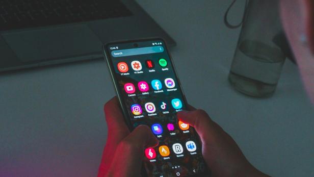 Person hält Smartphone mit geöffneten Social-Media-Apps vor einem Laptop auf einem Tisch.